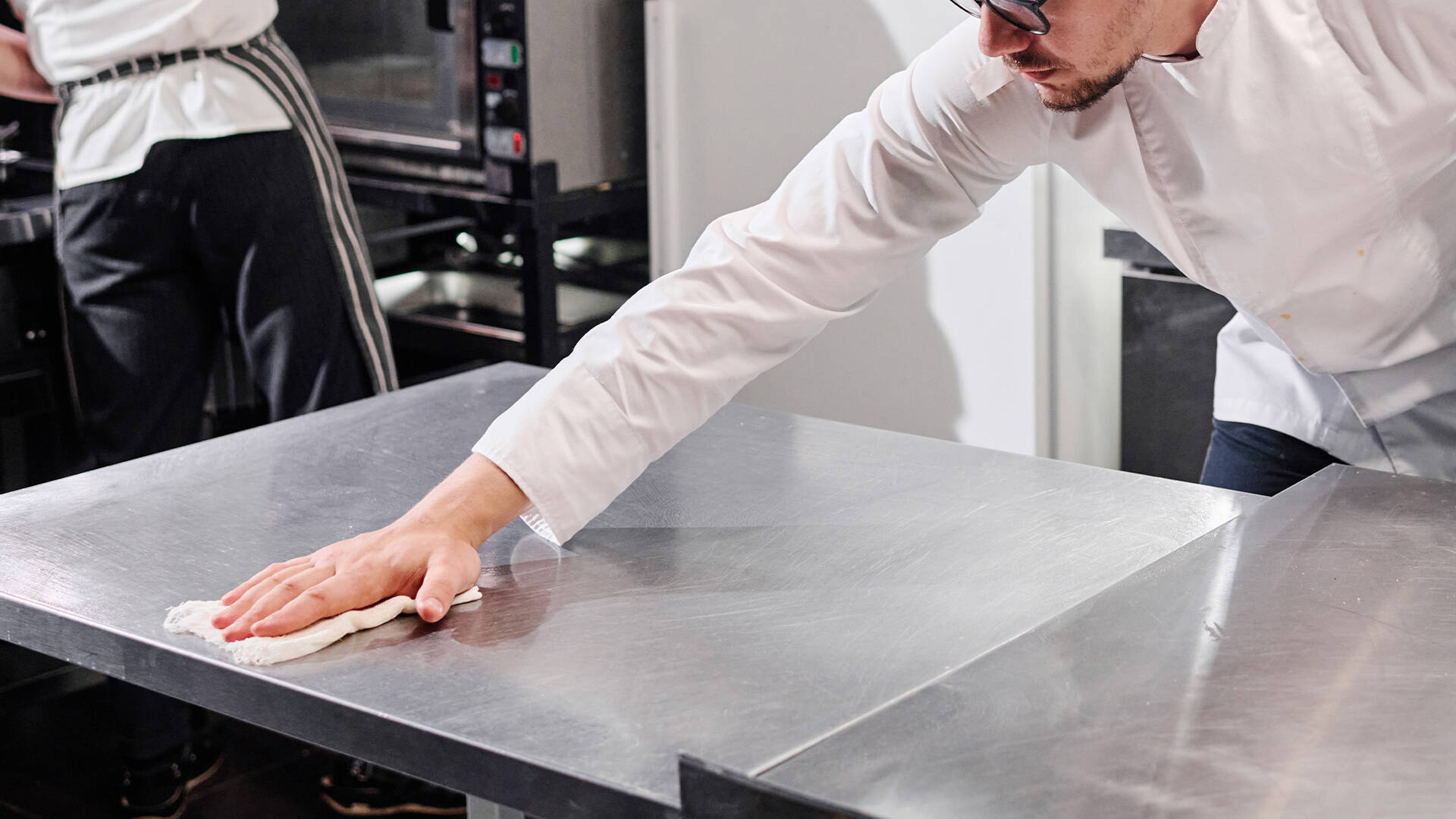 Chef wiping a stainless steel prep table with a disposable wiping cloth in a commercial kitchen
