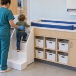 Caregiver in blue gloves helping a toddler climb the stairs of a daycare diaper changing station with a built-in sink